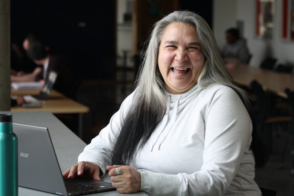 Indigenous student from the Fort McKay First Nation laughing in front of a computer in a campus library.