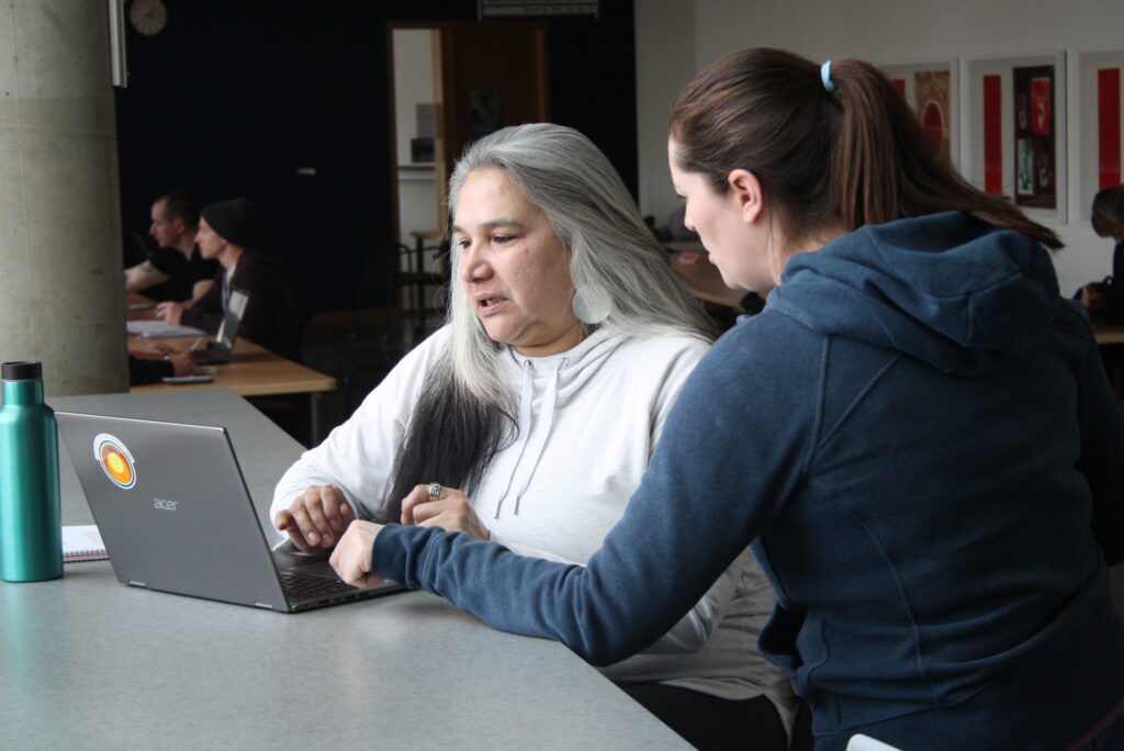 Two Indigenous students from the Metis and Fort McKay First Nations talking and studying in front of a laptop in a campus common space.