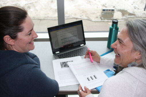 Two indigenous students from the Metis and Fort McKay First Nations in front of a laptop laughing and studying at a common space on campus.