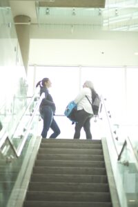 Two Indigenous students from the Metis and Fort McKay First Nations talking at the top of a staircase in a campus common space.