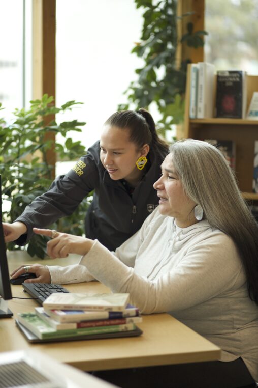 Two Indigenous students from the Stat'imc/Nlaka'pamux and Fort McKay First Nations studying using a computer in a campus library. Both students are pointing at the computer screen.