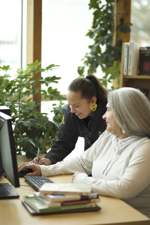 Two Indigenous students from the Stat'imc/Nlaka'pamux and Fort McKay First Nations studying by taking notes while sitting at a computer in a campus library.