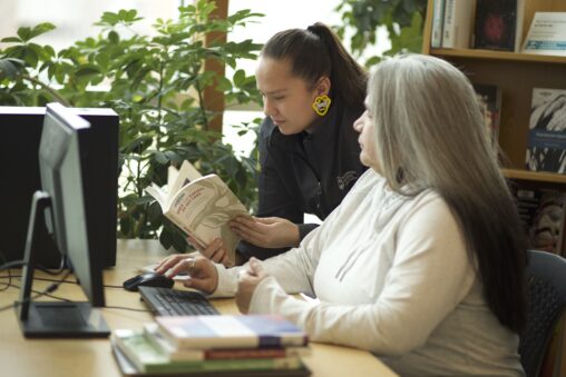 Two Indigenous students from the Stat'imc/Nlaka'pamux and Fort McKay First Nations reading a book in front of a computer in a campus library.