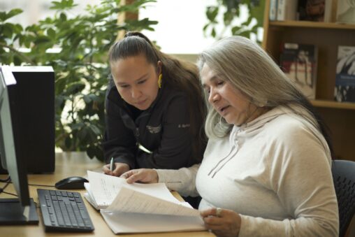 Two Indigenous students from the Stat'imc/Nlaka'pamux and Fort McKay First Nations studying by reading a notebook in front of a computer in a campus library.