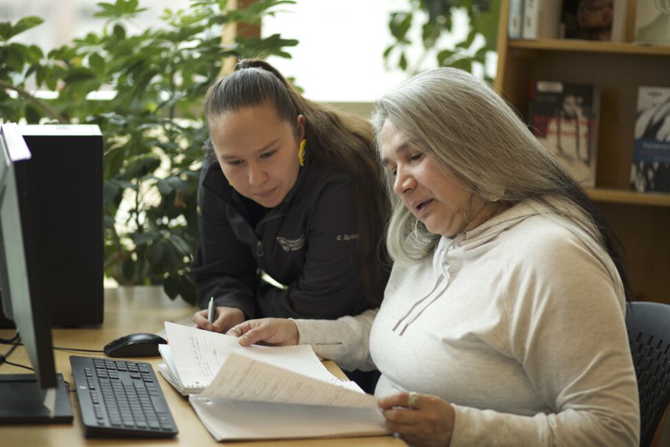 Two Indigenous students from the Stat'imc/Nlaka'pamux and Fort McKay First Nations studying by reading a notebook in front of a computer in a campus library.