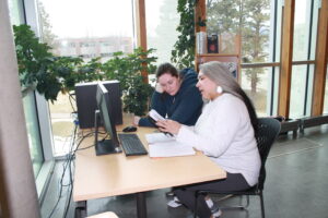 Two indigenous students from the Metis and Fort McKay First Nations studying in front of a computer work station at a campus library. One student is reading a book and talking to the other student.