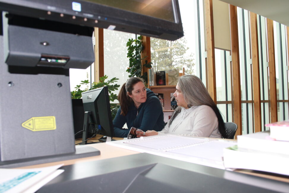 Two Indigenous students from the Metis and Fort McKay First Nations flipping through a book in front of a computer in a campus library.