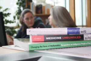 Three books about Indigenous people in the foreground and two Indigenous students from the Metis and Fort McKay First Nations studying at a campus library in the background.