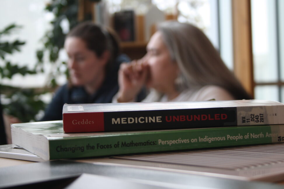 Two books about Indigenous people sit in the foreground while two Indigenous students from the Metis and Fort McKay First Nations are studying in the background in a campus library.