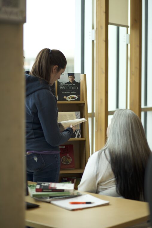 Two Indigenous students studying at a library on campus. One of the students is reading a book while the other is sitting and looking at the reading student.