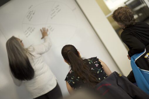 Indigenous student from the Fort McKay First Nation tutoring students from the St'at'imc/Nlaka'pamux Nation in front of a whiteboard.