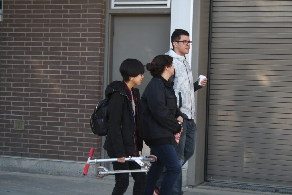 Three Indigenous students from the Haida and Ntekepmx Nations walking outdoors on campus. From left to right, they are a holding a paper coffee cup, a reusable water bottle, and a scooter, respectively.