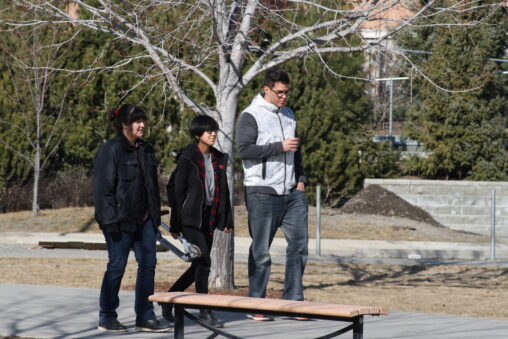 Three indigenous students from the Haida and Ntekepmx Nations walking on campus outdoors. The student in the middle carries a scooter.