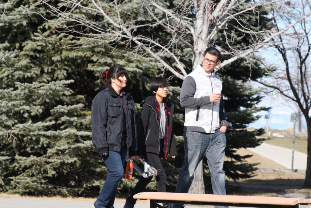 Three Indigenous students from the Haida and Ntekepmx Nations walking on campus outdoors. From left to right, each student is holding a reusable water bottle, a scooter, and a paper coffee cup, respectively.