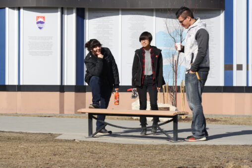 Three indigenous students from the Haida and Ntekepmx Nations hanging out on campus outdoors. The student in right has a paper coffee cup on their hand, the one in the middle carries a scooter and the student on the left has their foot on a bench and it holding a reusable water bottle.