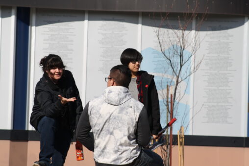 Three indigenous students from the Haida and Ntekepmx Nations on campus outdoors. Two students are standing up while the third one is sitting down on a bench.