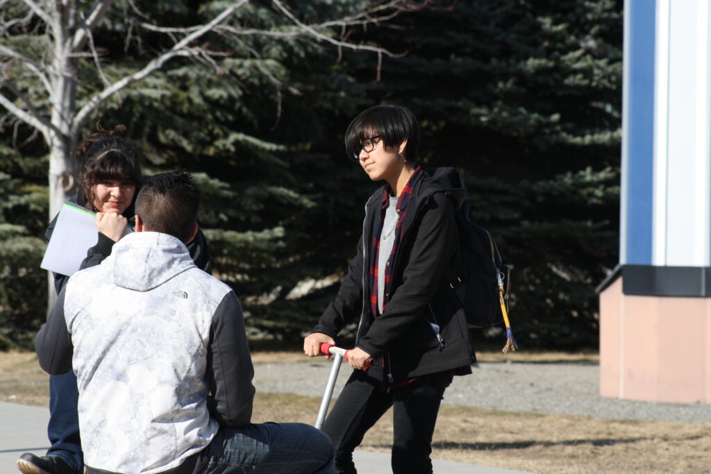 Three indigenous students from the Haida and Ntekepmx Nations on campus outdoors. Two students are standing up while the third one is sitting down on a bench.