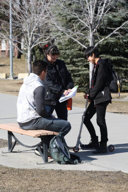 Three Indigenous students from the Haida and Ntekepmx Nations outdoors on campus. Two students are standing while the third one is sitting on a bench while looking at the notebook one of the standing students is showing them.