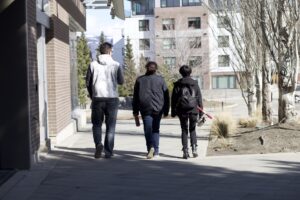 Three indigenous students from the Haida and Ntekepmx Nations walking outdoors on campus. Starting from the left, the first student is holding a coffee paper cup, the second a reusable water bottle and the third one a scooter.