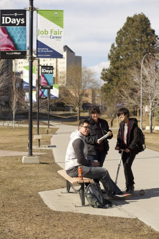 Three Indigenous students from the Haida and Ntekepmx Nations talking outside on campus. Two students are standing while the third one is sitting on a bench.