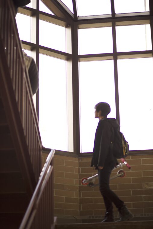 Indigenous student from the Ntekepmx Nation holding a scooter in one hand while climbing up the stairs inside a campus building.