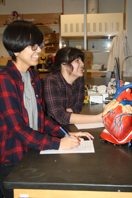 Two Indigenous students from the Ntekepmx Nation smiling while taking notes on a table that has a heart prop on it in an anatomy laboratory.