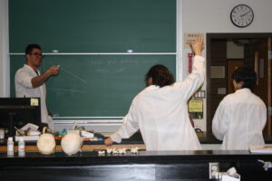 Three Indigenous students from the Haida and Ntekepmx Nations standing in front of a chalkboard in a science laboratory. One student is raising their hand to answer a question.