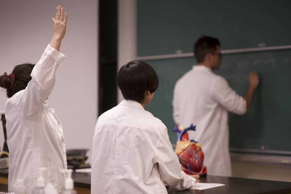 Indigenous student from the Haida Nation is peer mentoring their classmates while writing on a chalkboard in a biology laboratory. The two Indigenous students being mentored are sitting at a table and one is raising their hand.