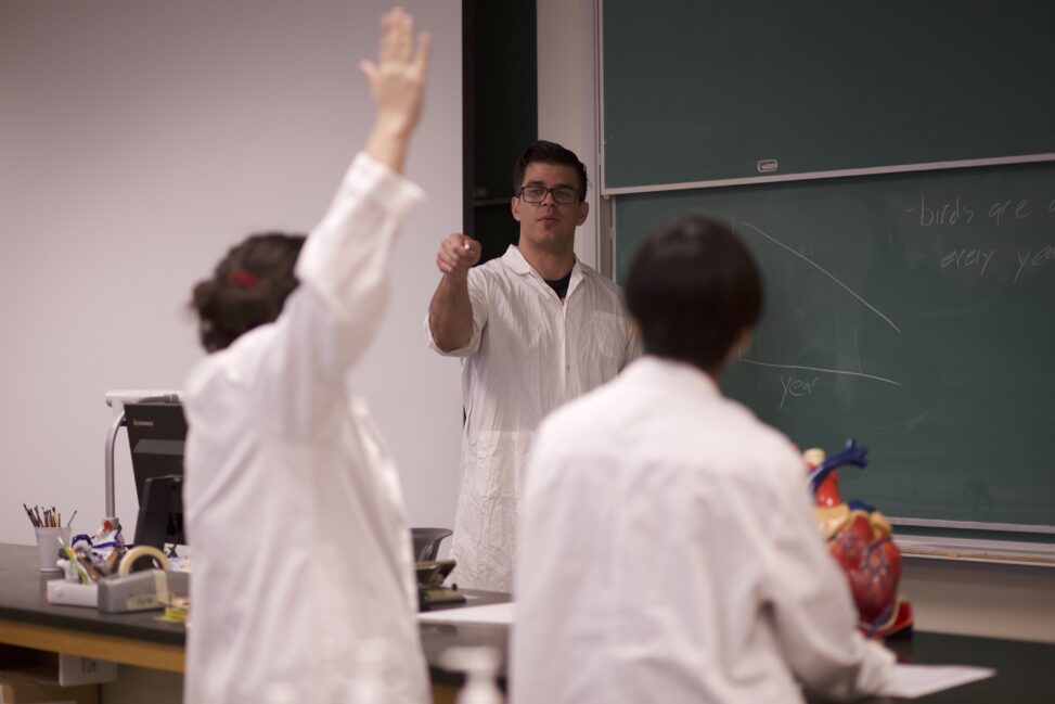 Three Indigenous students from the Haida and Ntekepmx Nations peer mentoring each other in a biology laboratory. One student is standing in front of chalkboard while the other two are sitting with one raising their hand.