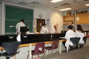 Four Indigenous students from the Haida, Ntekepmx and Metis Nations tutoring peers in a biology laboratory. Two students are standing in front of a chalkboard. The other two are sitting and listening.