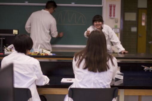 Four Indigenous students from the Haida, Ntekepmx and Metis Nations mentoring each other in a biology laboratory. Two students are in front of the chalkboard while the other two are sitting and listening.