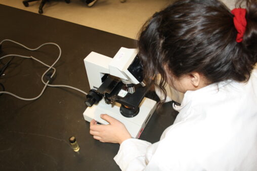 Indigenous student from the Ntekepmx Nation using a microscope in a science/biology laboratory. A small jar containing a liquid sample sits beside the microscope.