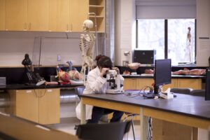 Indigenous student from the Ntekepmx Nation looking through a microscope in a biology laboratory. Multiple anatomy props are in the background.