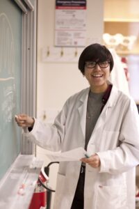 Indigenous student from the Ntekepmx Nation smiling at the camera while pointing a piece of chalk at the chalkboard beside them.