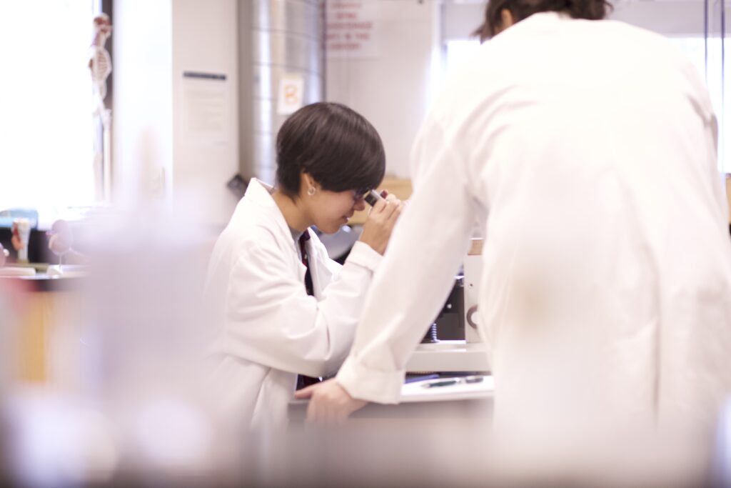 Indigenous students from the Ntekepmx Nation using a microscope in a biology laboratory. One of the students is looking through the microscope while the other is leaning on the table beside them.