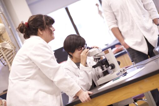 Indigenous students from the Ntekepmx Nation in a biology lab. One of the students is looking through the microscope while the other one is waiting for their turn.