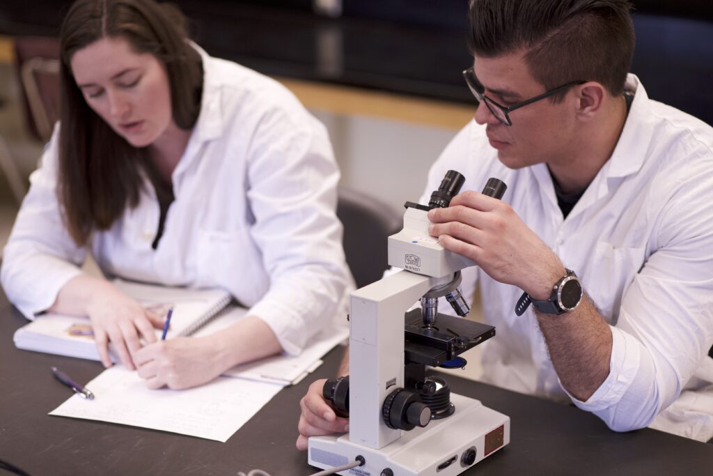 Two Indigenous students from the Metis and Haida Nations in a biology laboratory. The student on the left is taking notes while the one on the right is sitting in front of a microscope and looking at the other student's notes.