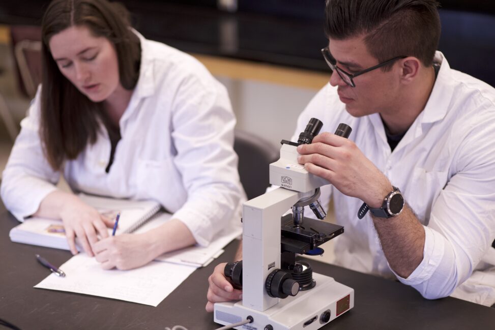 Two Indigenous students from the Metis and Haida Nations in a biology laboratory. The student on the left is taking notes while the one on the right is sitting in front of a microscope and looking at the other student's notes.