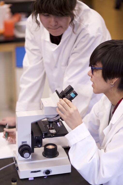 Two Indigenous students from the Ntekepmx Nation with a microscope in a biology laboratory. One student is using the microscope while the other is leaning on the table beside them.