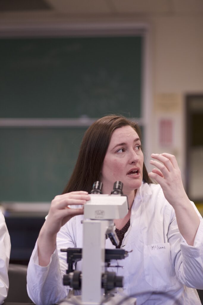 Indigenous student sitting in front of a microscope and explaining something to someone off camera on the right in a biology laboratory.