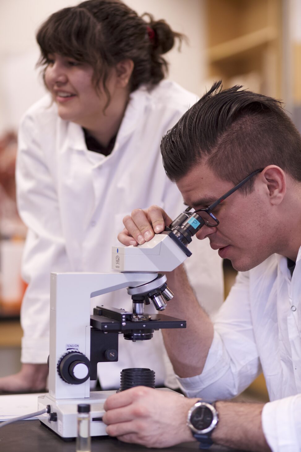 Indigenous students from the Haida and Ntekepmx Nations using a microscope in a biology laboratory. One student is looking through the microscope while the other is waiting for their turn.