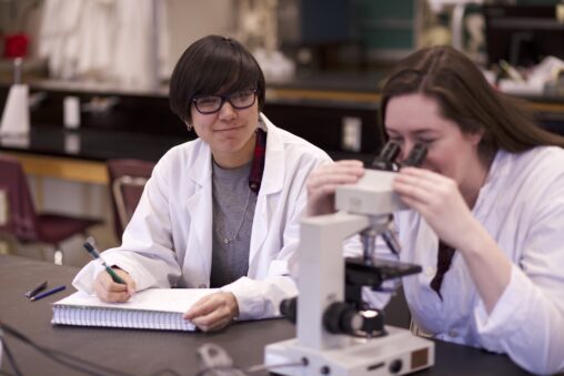 Two Indigenous students from the Metis and Ntekepmx Nations using a microscope in a biology laboratory. One student is using the microscope while the other is taking notes.