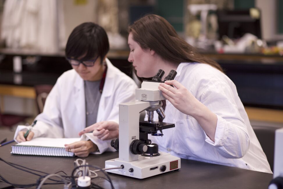 Two Indigenous students from the Metis and Ntekepmx Nations in a biology laboratory. The student on the left is taking some notes, and the student on the right is holding the microscope with one hand while talking with the other student.