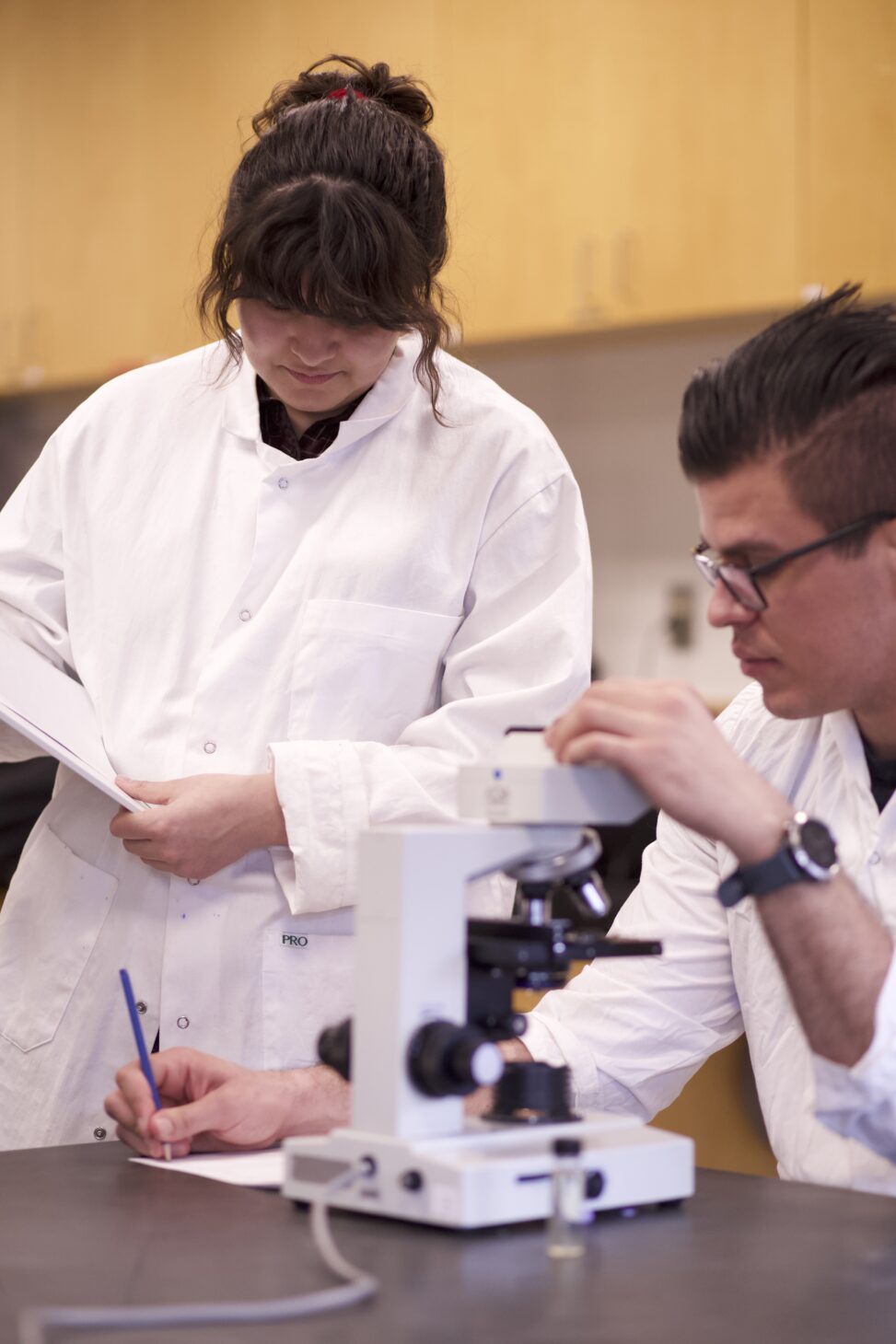 Two Indigenous students from the Haida and Ntekepmx Nations in a biology lab. The student on the left is looking at the notes that the student on the write is taking while resting their other hand on a microscope.