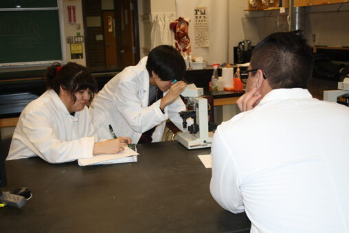 Three Indigenous students from the Haida and Ntekepmx Nations in a biology lab. The student on the left is taking notes, the student in the center is looking through a microscope, and the student on the right is watching the other two.