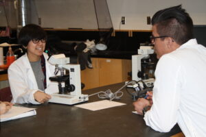 Two Indigenous students from the Haida and Ntekepmx Nations sitting with two microscopes in a biology laboratory.