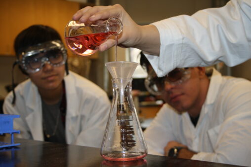 Two Indigenous students from the Haida and Ntekepmx Nations watching another student pouring liquid from one Erlenmeyer flask into another in a chemistry laboratory.