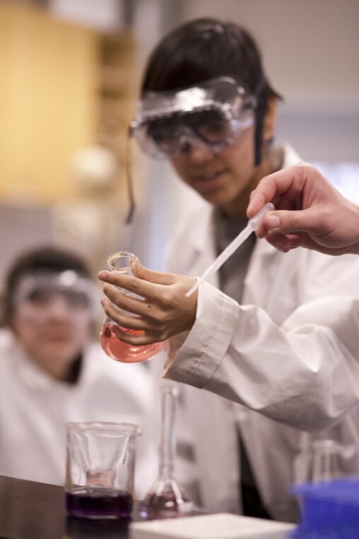 Indigenous student grabbing an Erlenmeyer flask from a table in a chemistry laboratory. In the foreground, a student's hand holds a dropper while, in the background, another student watches.