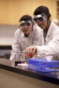 Two Indigenous students from the Ntekepmx Nation in a chemistry laboratory. The student on the left is watching the student on the right pour liquid from an Erlenmeyer flask into a beaker.