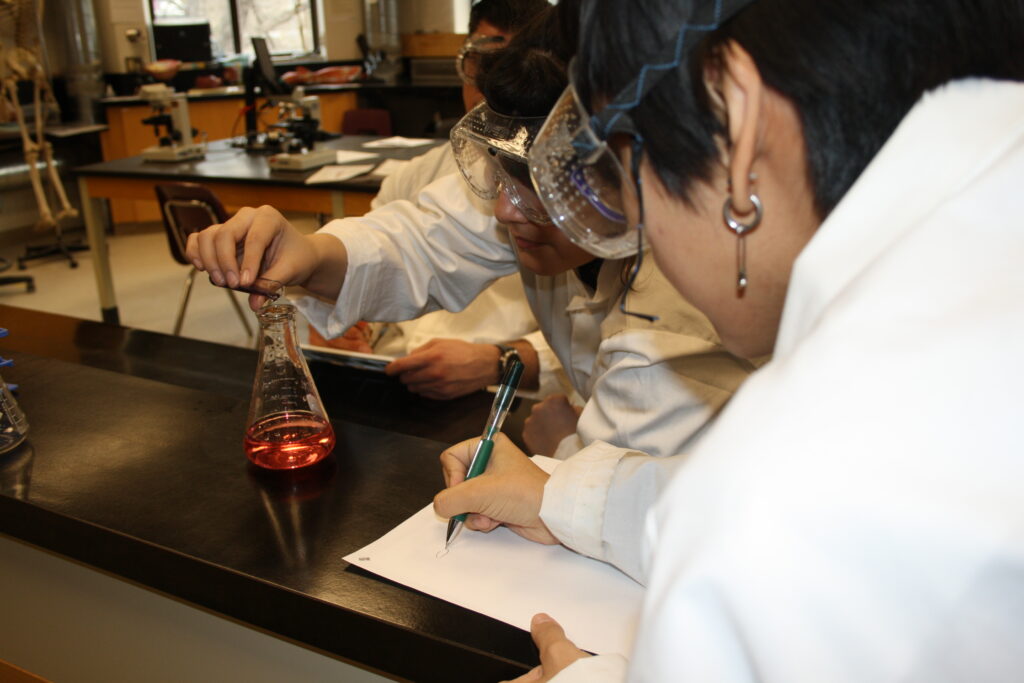 Two Indigenous students from the Ntekepmx Nation conducting an experiment by having one student pour liquid into an Erlenmeyer flask while the other takes notes in a chemistry laboratory.